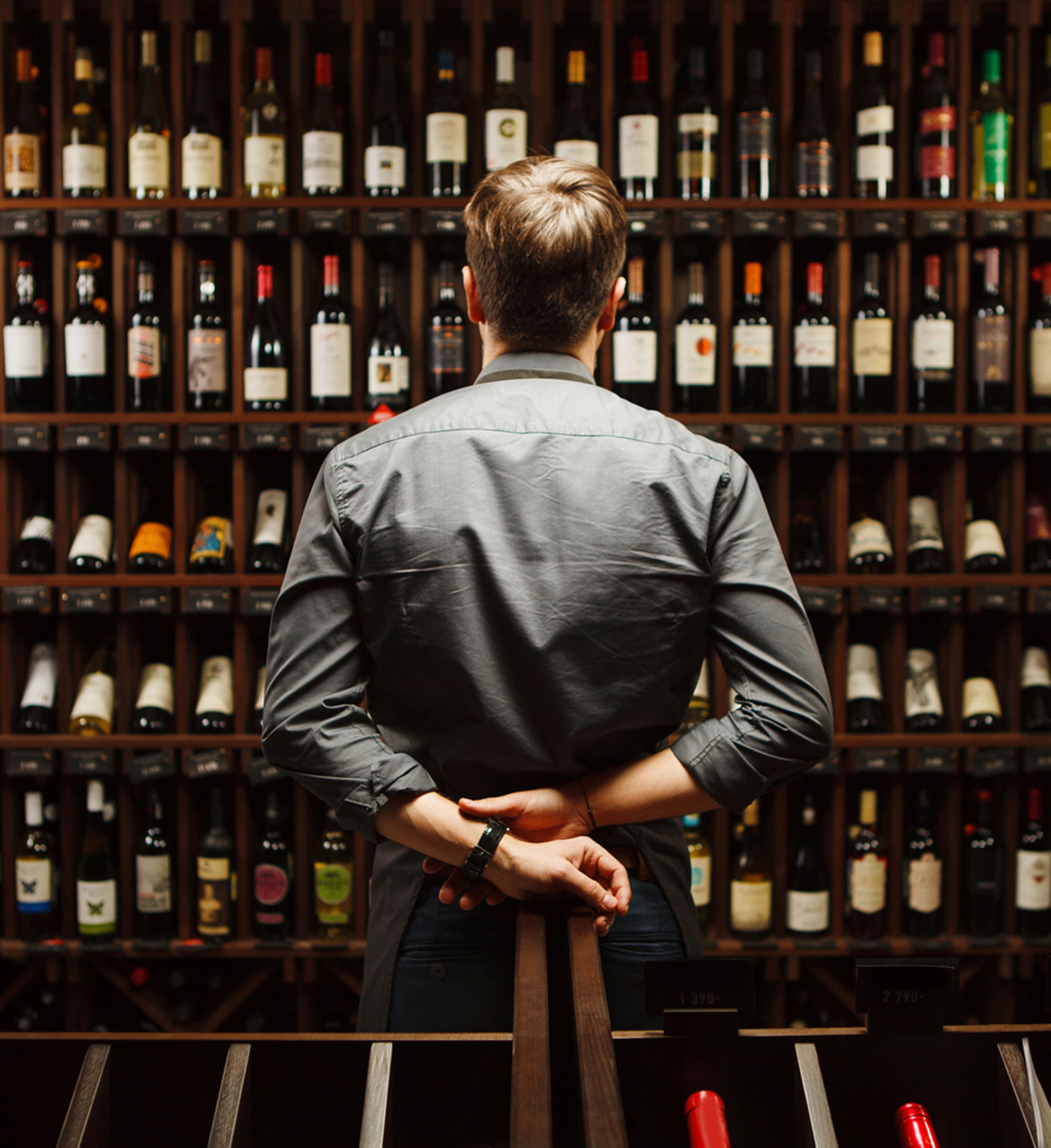 Bartender at wine cellar full of bottles with exquisite alcohol drinks that have various sweet and sour tastes and dates of manufacture on large wooden shelves.